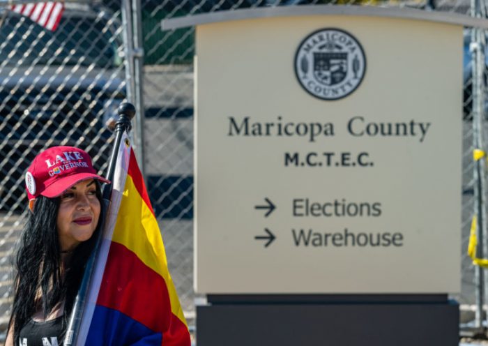 A right wing activist wearing a 'LAKE FOR GOVERNOR' hat stands on the sidewalk in protest of the election process in front of the Maricopa County Tabulation and Election Center on November 14, 2022 in Phoenix, Arizona. Ballots continue to be counted in Maricopa County following the November 8 midterm election as officials push back against conspiracy theories claiming the process is being delayed. (Photo by Jon Cherry/Getty Images)