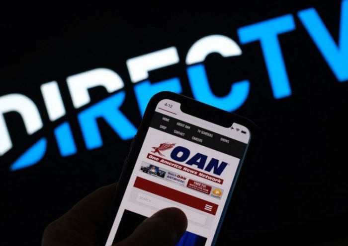 This illustration photo shows a person reading the One America Network website on a smartphone in front of a DirecTV logo in Los Angeles, January 14, 2022. - Subscription television service DirecTV has decided not to renew its contract with One America News Network (OAN), an ultra-conservative, conspiratorial US channel that backs former US president Donald Trump."We informed (OAN owner) Herring Networks that, following a routine internal review, we do not plan to enter into a new contract when our current agreement expires," a spokesman for DirecTV told AFP January 14, 2021. (Photo by Chris DELMAS / AFP) (Photo by CHRIS DELMAS/AFP via Getty Images)