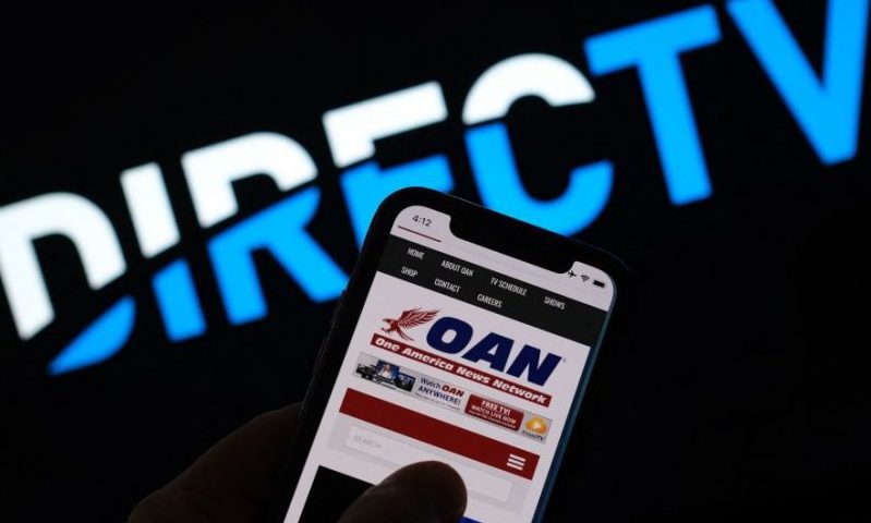 This illustration photo shows a person reading the One America Network website on a smartphone in front of a DirecTV logo in Los Angeles, January 14, 2022. - Subscription television service DirecTV has decided not to renew its contract with One America News Network (OAN), an ultra-conservative, conspiratorial US channel that backs former US president Donald Trump."We informed (OAN owner) Herring Networks that, following a routine internal review, we do not plan to enter into a new contract when our current agreement expires," a spokesman for DirecTV told AFP January 14, 2021. (Photo by Chris DELMAS / AFP) (Photo by CHRIS DELMAS/AFP via Getty Images)