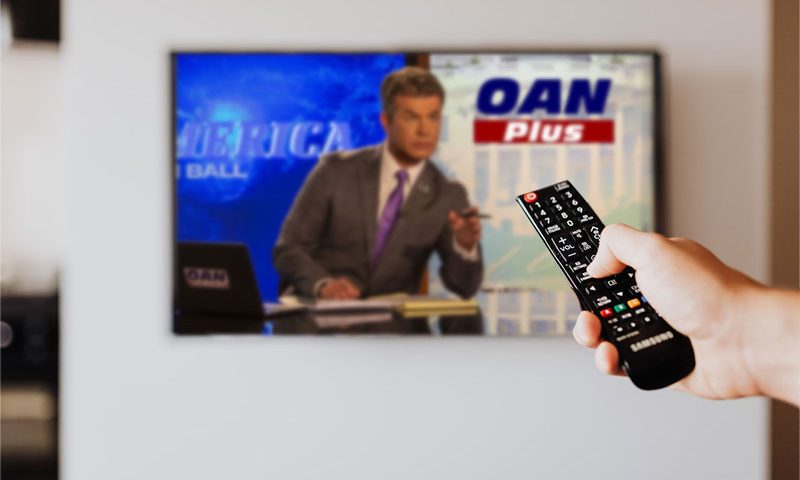 Person pressing the button of a remote control with One America News on the television in the background. Photo by Karolina Grabowska