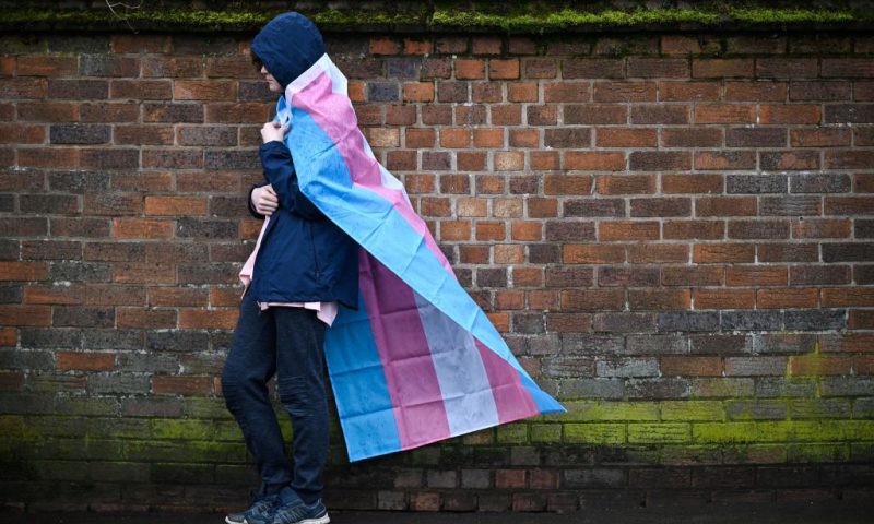 A mourner wrapped in the Transgender Flag waits for the horse-drawn carriage transporting the coffin of murdered transgender teenager Brianna Ghey, outside St Elphin's Church in Warrington, northern England on March 15, 2023, at the end of the funeral service. - 16-year-old Brianna Ghey's body was discovered by members of the public in a park in Culcheth, north of Warrington, after having been fatally stabbed. (Photo by Oli SCARFF / AFP) (Photo by OLI SCARFF/AFP via Getty Images)