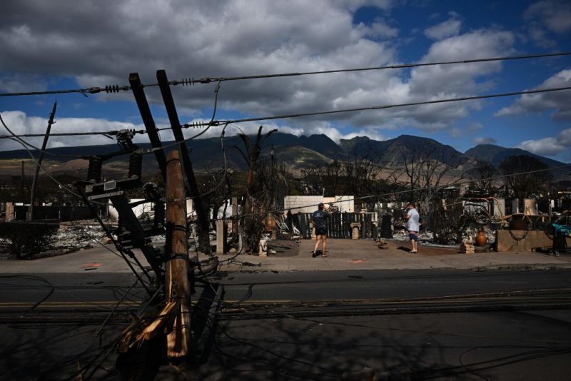 Downed power lines block a road as people feed chickens outside a burnt home in the aftermath of a wildfire in Lahaina, western Maui, Hawaii on August 11, 2023. A wildfire that left Lahaina in charred ruins has killed at least 67 people, authorities said on August 11, making it one of the deadliest disasters in the US state's history. Brushfires on Maui, fueled by high winds from Hurricane Dora passing to the south of Hawaii, broke out August 8 and rapidly engulfed Lahaina. (Photo by Patrick T. Fallon / AFP) (Photo by PATRICK T. FALLON/AFP via Getty Images)