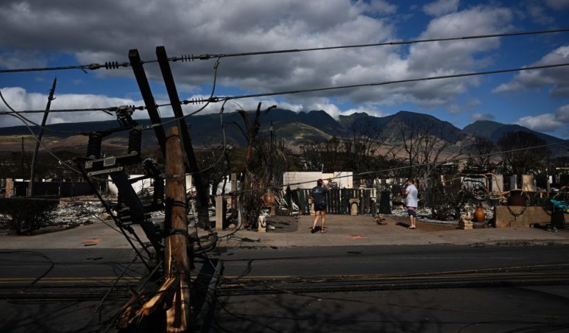 Downed power lines block a road as people feed chickens outside a burnt home in the aftermath of a wildfire in Lahaina, western Maui, Hawaii on August 11, 2023. A wildfire that left Lahaina in charred ruins has killed at least 67 people, authorities said on August 11, making it one of the deadliest disasters in the US state's history. Brushfires on Maui, fueled by high winds from Hurricane Dora passing to the south of Hawaii, broke out August 8 and rapidly engulfed Lahaina. (Photo by Patrick T. Fallon / AFP) (Photo by PATRICK T. FALLON/AFP via Getty Images)