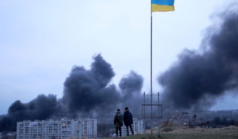 People standing near a Ukrainian national flag watch as dark smoke billows following an air strike in the western Ukrainian city of Lviv, on March 26, 2022. At least five people wounded in two strikes on Lviv, the regional governor said, in a rare attack on a city that has escaped serious fighting since Russian troops invaded last month. (Photo by Oleksii FILIPPOV / AFP) (Photo by OLEKSII FILIPPOV/AFP via Getty Images)