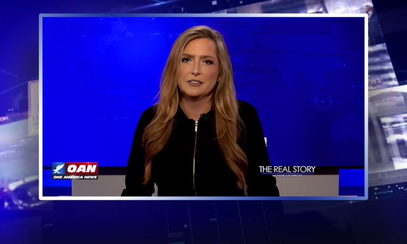 Video still of the host of The Real Story at the desk of their talk show on One America News Network.