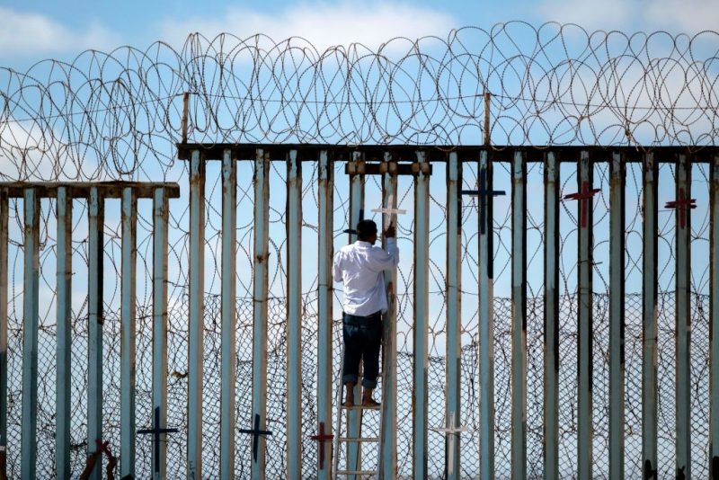 TOPSHOT - Josue Serrano, a Mexican deported migrant, hangs wooden crosses on the border fence as part of a vigil for migrants who died while migrating to the United States, at the US-Mexico border in playas de Tijuana, Baja California state, Mexico, on July 4, 2022. - On June 27, 2022, 53 migrants from Mexico, Guatemala and Honduras were found death abandoned in a suffocatingly hot tractor-trailer truck in the US state of Texas. (Photo by Guillermo Arias / AFP) (Photo by GUILLERMO ARIAS/AFP via Getty Images)