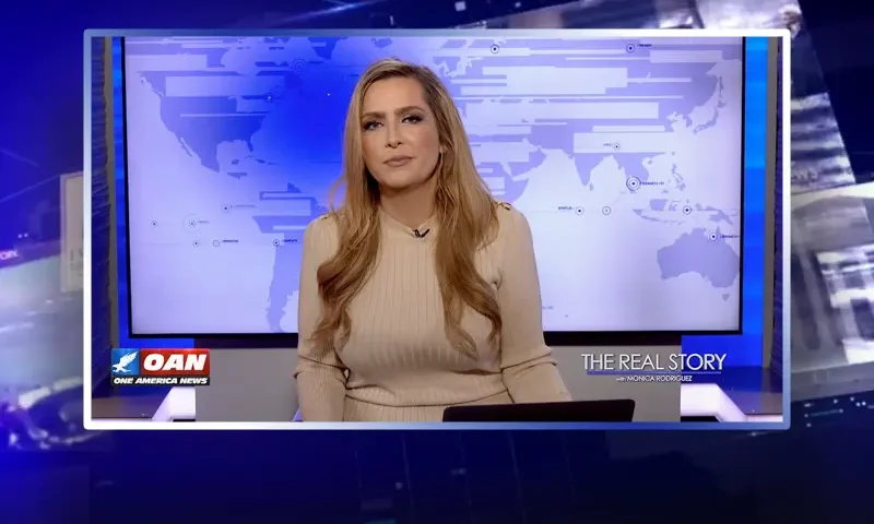 Video still of the host of The Real Story at the desk of their talk show on One America News Network.