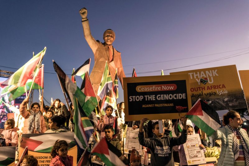 People raise flags and placards as they gather around a statue of late South African president Nelson Mandela to celebrate a landmark "genocide" case filed by South Africa against Israel at the International Court of Justice, in the occupied West Bank city of Ramallah on January 10, 2024. The ruling African National Congress (ANC) has long been a firm supporter of the Palestinian cause, often linking it to its own struggle against the white-minority government, which had cooperative relations with Israel. (Photo by MARCO LONGARI / AFP) (Photo by MARCO LONGARI/AFP via Getty Images)