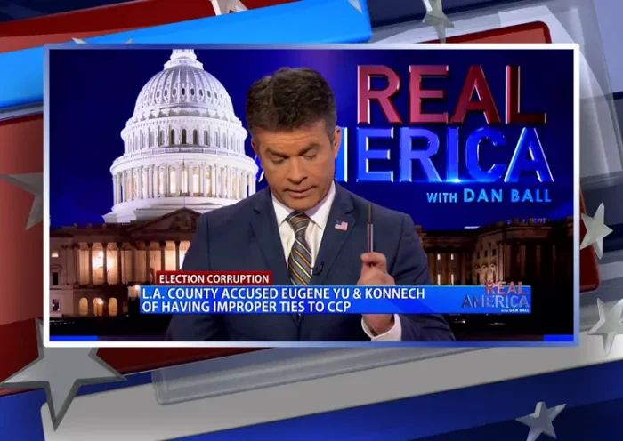 Video still of the host of Real America at the desk of their talk show on One America News Network.